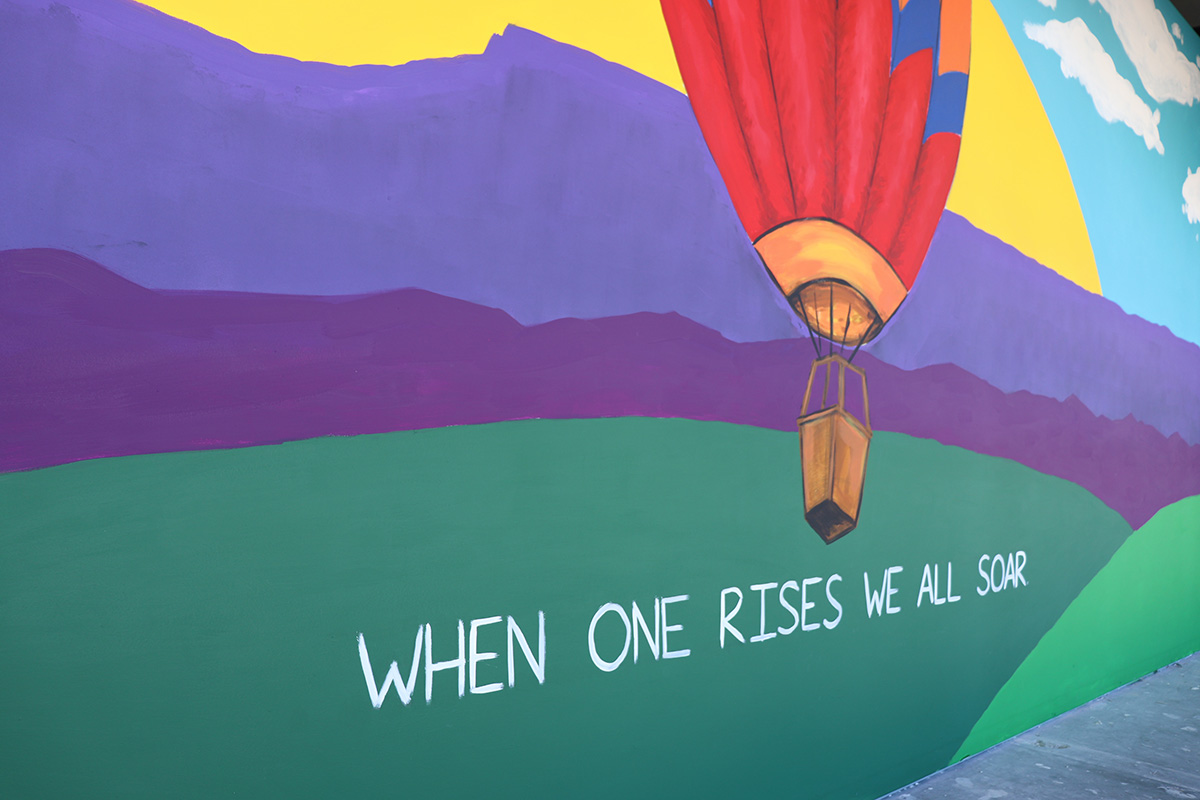 A mural depicting a colorful hot air balloon rises with the sunset in the background. White text under the hot air balloon reads "When One Rises, We All Soar".