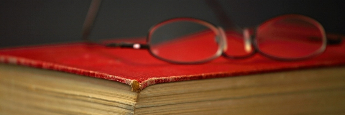 Older book with red cover and reading glasses lying upon it. 