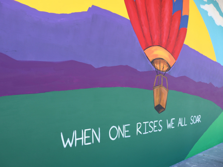 A mural depicting a colorful hot air balloon rises with the sunset in the background. White text under the hot air balloon reads "When One Rises, We All Soar".