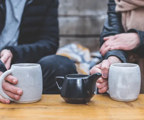 Close-up image of two coffee mugs being held by individuals. A pitcher of creamer rests on the table between the two cups.