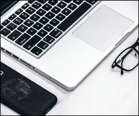 Keyboard laptop. To the left of the laptop rests a cell phone. A pair of eye glasses are folded up and resting in front of the laptop. 