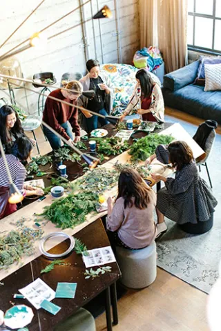 Group of women standing around a table working on crafts with lots of greenery