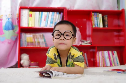 Young boy wearing yellow shirt and glasses sitting at table with red bookshelves in the background