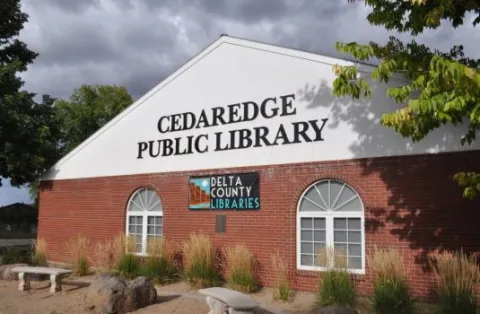 Brick and white building with "Cedaredge Public Library" sign against a cloudy sky